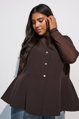 Kvinde med langt mørkt hår iført en mørkebrun peplum-top med knapper og lyseblå jeans, rører ved sit hår og kigger ned.