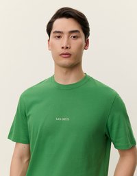 Groene katoenen T-shirt met ronde hals en korte mouwen. De voorkant heeft witte geborduurde tekst met de woorden "LES DEUX." Soepele textuur.