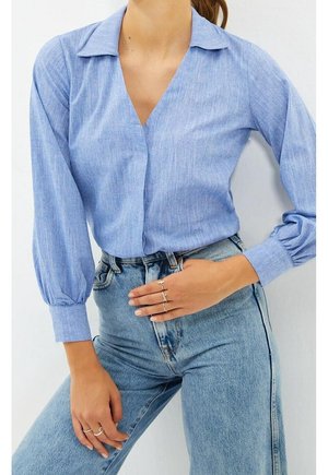 Frau trägt ein hellblaues Langarmshirt mit V-Ausschnitt und hoch taillierte hellblaue Jeans, zeigt Ringe an beiden Händen.