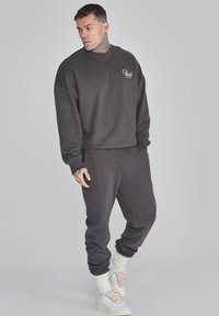 Conjunto de sudadera y pantalones de chándal en gris oscuro, hecho de un tejido suave. Presenta un corte relajado, puños acanalados y un pequeño logo en el pecho.