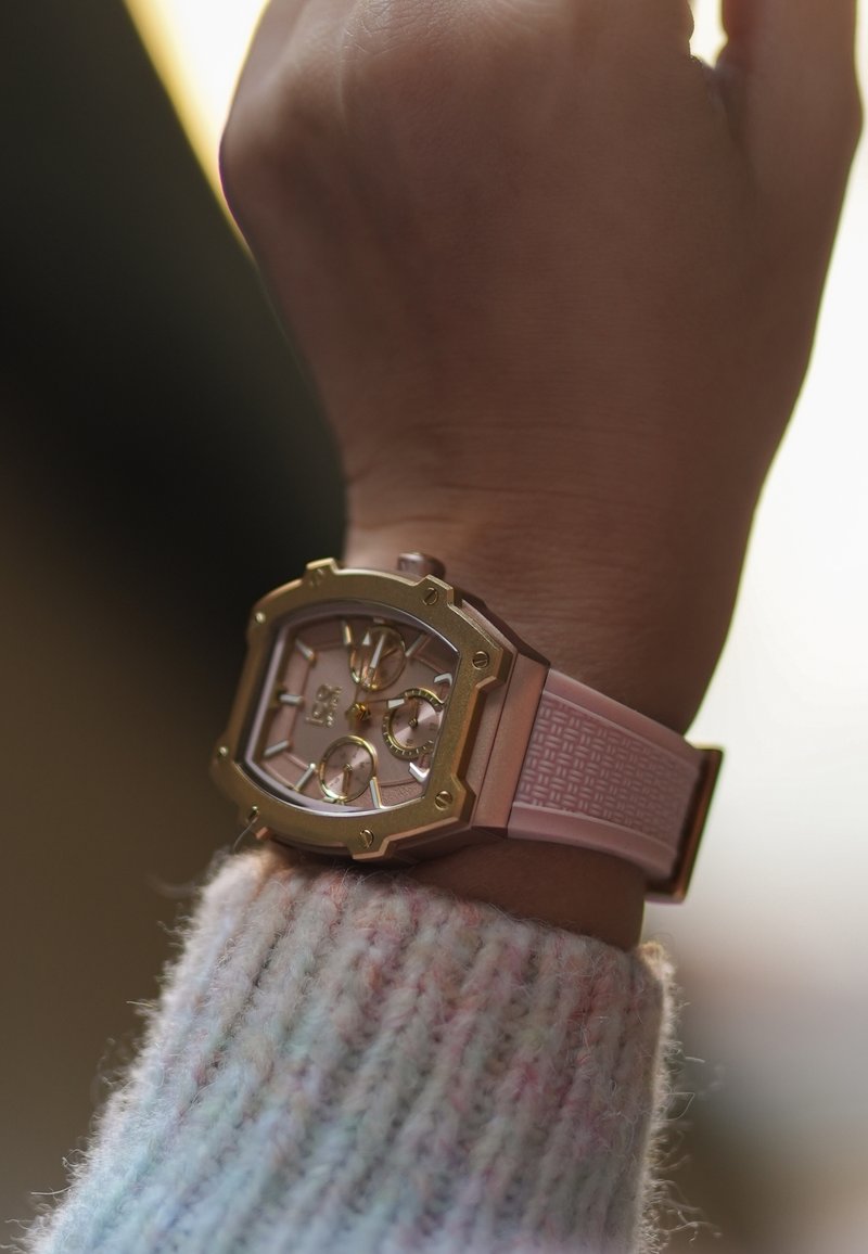 Montre rectangulaire en or rose avec trois sous-cadrans sur un bracelet en caoutchouc texturé rose clair. Portée sur une main avec un pull en tricot.