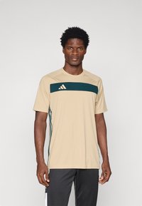 Beżowa koszulka sportowa z ciemnozielonym poziomym paskiem, logo Adidas na lewej piersi, krótkie rękawy, okrągły dekolt, materiał odprowadzający wilgoć.