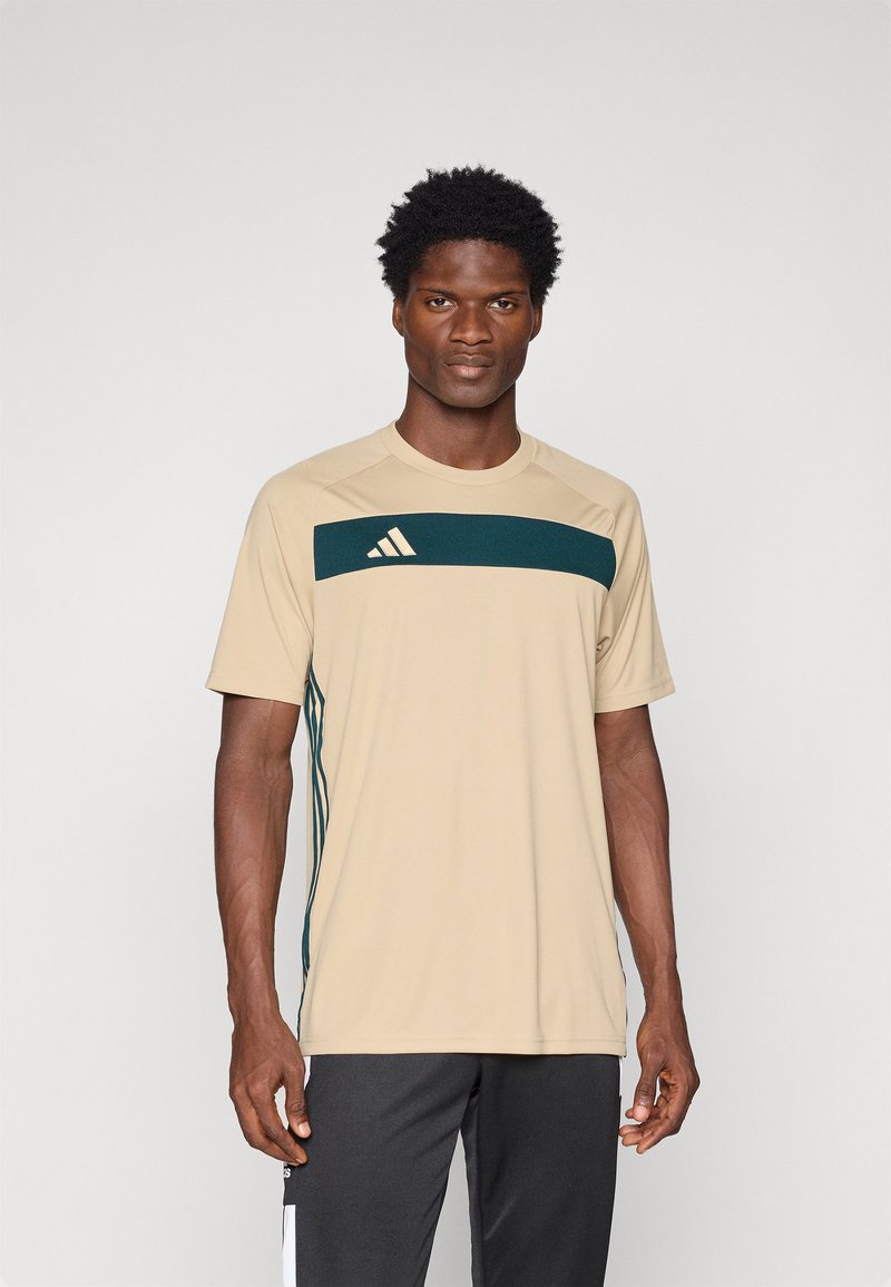 Beżowa koszulka sportowa z ciemnozielonym poziomym paskiem, logo Adidas na lewej piersi, krótkie rękawy, okrągły dekolt, materiał odprowadzający wilgoć.