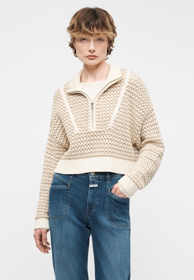 Νεαρός με κοντά μαλλιά που φοράει μπεζ πλεκτό πουλόβερ με φερμουάρ στη μέση και μπλε τζιν, στέκεται με το ένα χέρι στην τσέπη.