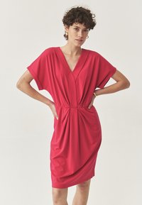 Rotes ärmelloses Kleid mit V-Ausschnitt, gerafften Details an der Taille und drapiertem Design, hergestellt aus einem glatten, leichten Stoff.