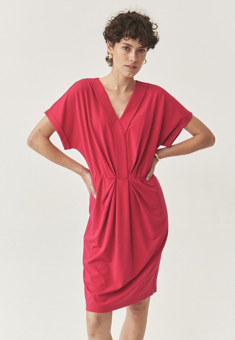 Rotes ärmelloses Kleid mit V-Ausschnitt, gerafften Details an der Taille und drapiertem Design, hergestellt aus einem glatten, leichten Stoff.