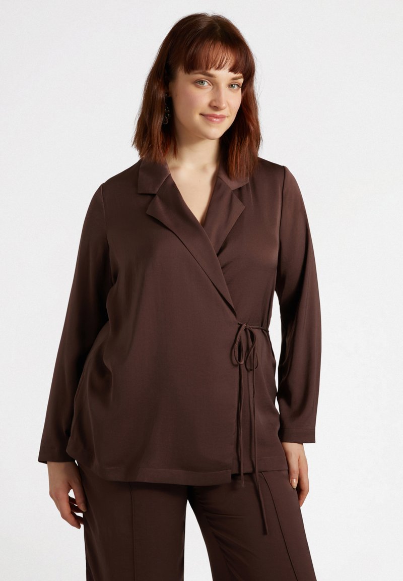 Brązowa bluzka z długim rękawem, zapinana na krzyż, z wyciętym kołnierzem, wiązaniem w talii i gładką fakturą. Połączona z pasującymi brązowymi spodniami.