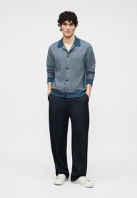 Mann som står med hendene i lommene, iført en blå mønstret knappet cardigan, mørke pinstripede bukser og hvite sneakers mot hvit bakgrunn.