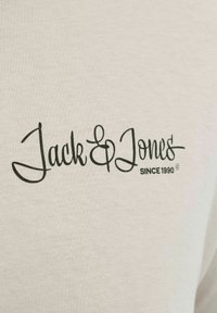 Jasnobeżowa bawełniana koszulka z wyraźnym czarnym logo w stylu pisma, z napisem "Jack & Jones" oraz "SINCE 1990." Gładka faktura, luźny krój.