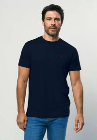 Jūras zila īsa piedurkņu T-kreklā no mīksta materiāla ar apaļu kaklu un mazu sarkanu logotipu kreisajā krūtī. Valkāts ar ziliem džinsiem.