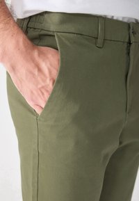 Männliche Hand, die in der vorderen Tasche einer olivgrünen Hose ruht, die sichtbare Gürtelschlaufen und einen Reißverschluss hat.