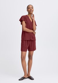 Completo due pezzi color bordeaux con top a scollatura a V e maniche a balza, abbinato a pantaloni corti. Tessuto morbido, vestibilità comoda. Scarpe marroni slip-on.