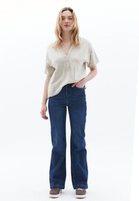 Beige Kurzarmoberteil mit V-Ausschnitt und Knopfleiste, kombiniert mit hochgeschnittenen dunkelblauen Schlagjeans und braunen Schuhen.