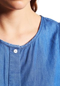 Kvinna som bär en blå denimtop med en liten vit knapp vid den runda halsringningen, som visar halsen och en del av det nedre ansiktet.