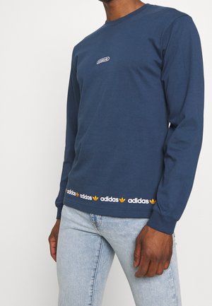 Mężczyzna ubrany w granatową, długorękawową koszulkę Adidas z małym logo na piersi oraz powtarzającym się napisem Adidas i logo koniczyny wzdłuż dolnego brzegu.