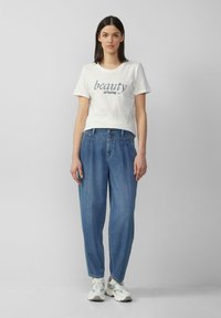 T-shirt en coton blanc avec "la beauté d'être" imprimé en bleu marine, associé à un jean en denim bleu clair à coupe ample et des baskets blanches.