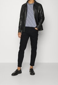 Uomo che indossa una giacca di pelle nera sopra una camicia a righe bianco e nero, pantaloni neri e mocassini neri, in piedi contro uno sfondo semplice.