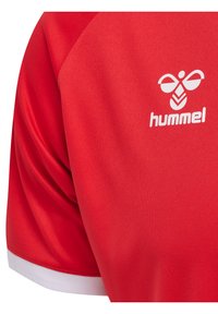 Röd sporttröja i slätt tyg. Har en vit accent vid kragen och en vit "hummel" logotyp på övre vänstra sidan.