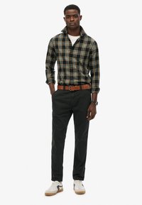 Chemise à carreaux bleu marine et jaune, pantalon sombre, ceinture marron et baskets blanches. Le modèle se tient avec les bras détendus le long du corps.