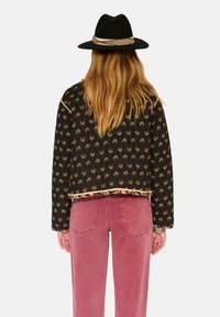 Veste noire à motifs floraux avec bordure dorée, coupe courte ; associée à un jean rose. Le mannequin porte un chapeau en feutre noir.