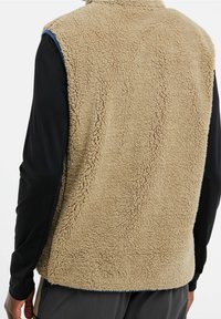 Personne portant un gilet polaire beige par-dessus une chemise noire à manches longues, vue de dos, mettant en valeur le tissu doux et texturé du gilet.