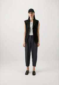 Giovane donna che indossa un cappello nero, un blazer senza maniche nero, una maglietta bianca, pantaloni navy a vita alta e mocassini neri, in piedi contro uno sfondo uniforme.