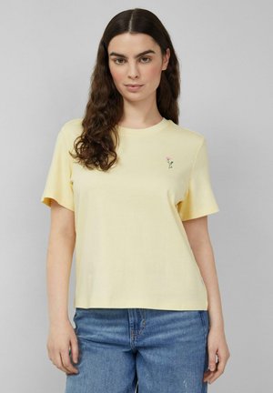 Frau mit langen, dunklen, lockigen Haaren, die ein hellgelbes Kurzarmshirt mit kleinem pinkfarbenem Blumendetail und blaue Jeans trägt.