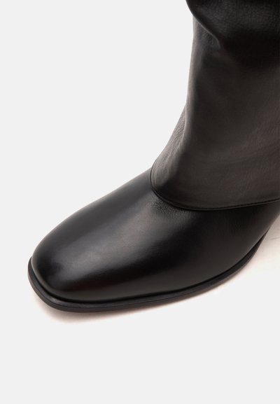 Botte en cuir noir avec un bout carré arrondi et une texture lisse, vue en gros plan sur un fond blanc uni.