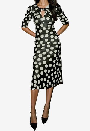 Femme portant une robe midi noire cintrée à gros pois blancs, manches trois-quarts, encolure avec ouverture en goutte d'eau, et escarpins noirs à bout pointu.