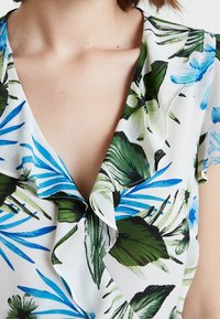 Blusa floreale in tessuto leggero con foglie e fiori verdi e blu su sfondo bianco, con dettagli di colletto arricciato.