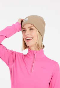 Beanie a coste color sabbia con un piccolo patch logo, abbinato a un top rosa a maniche lunghe con zip a un quarto e una trama liscia e elastica.