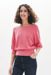 Maglione rosa a maglia con scollo a barca, maniche a tre quarti e polsini a costine. Abbinato a jeans blu scuro, con tasca laterale.