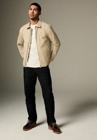Veste légère beige avec un col, T-shirt blanc, jeans foncés et chaussures à lacets marron avec semelles en caoutchouc. Design simple et fonctionnel.