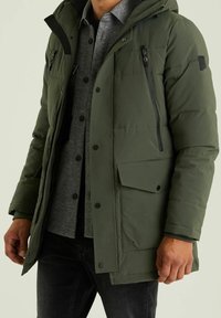 Parka imbottito di colore verde scuro, con chiusura frontale a cerniera, multiple tasche e cappuccio. Indossato sopra una camicia grigia a motivo e jeans scuri.
