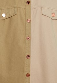 Gros plan sur le devant d'une chemise avec deux poches poitrine en tissu beige et beige clair, avec des boutons-pression couleur cuivre au centre.