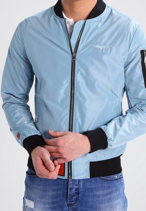 Mann trägt eine hellblaue Bomberjacke mit schwarzem Kragen und Bündchen, die zugezippt ist, kombiniert mit ausgewaschenen blauen Jeans.