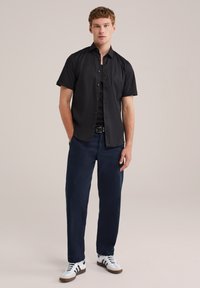 Young man standing with hands in pockets wearing a black short-sleeve button-up shirt, dark blue pants, and white sneakers with black stripes.