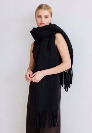 Femme blonde portant un haut sans manches noir et un pantalon sombre,.accessoirisée d'une grande écharpe noire à franges enroulée autour du cou.