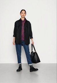 Camisa oversized preta, blusa com padrão roxo, jeans azul com mangas enroladas, botas pretas de tornozelo e uma grande bolsa preta. Fundo simples.