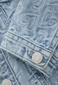 Denimkangas hele sinise pesuga, millel on tikitud lillemustrid ja metallist nupud, millele on graveeritud "Next Essential" jope mansetil.
