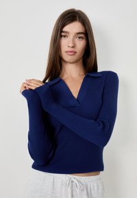 Młoda kobieta z długimi brązowymi włosami, ubrana w granatowy sweter z prążkami i jasnoszare spodnie, stojąca na tle białej ściany.