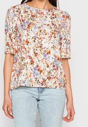 Kvinde iført en kortærmet hvid bluse med flerfarvet blomsterprint kombineret med lyseblå jeans.