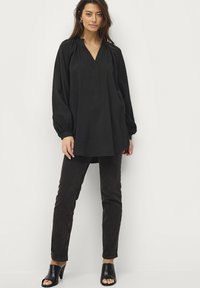 Sort oversized bluse med lange ærmer og V-udskæring, struktureret stof og samlede skuldre, parret med mørkebrune slim-fit jeans og sorte åbne hæle.