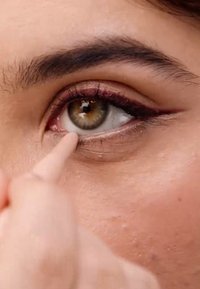 Gros plan sur un œil marron clair avec un eyeliner noir. Un doigt applique un produit sur la peau de la paupière inférieure. Texture de la peau lisse visible.
