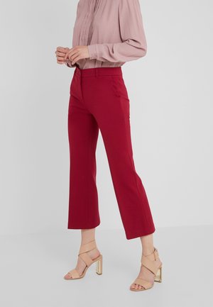 Mujer que lleva una blusa rosa de manga larga, pantalones rojos recortados y sandalias beige de tacón alto, de pie contra un fondo claro y liso.