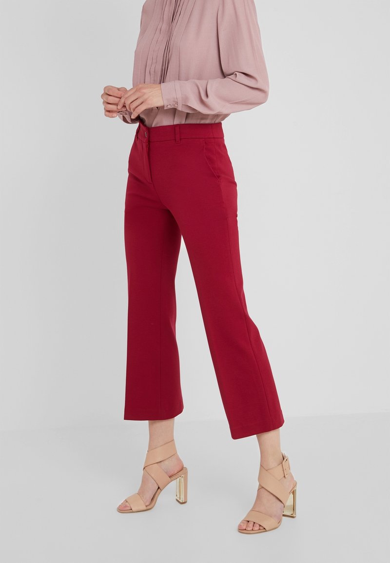 Femme portant un chemisier rose à manches longues, un pantalon rouge raccourci et des sandales beiges à talons hauts, debout devant un fond clair uni.
