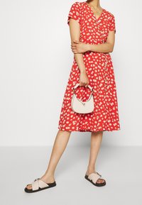 Vestido rojo con estampado floral blanco, de mangas cortas, diseño envolvente y falda acampanada. Combinado con un bolso rosa claro y sandalias negras tipo slide.