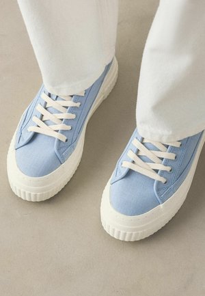 Lyseblå lærreds-sneakers med tykke hvide gummisåler og hvide snørebånd, båret med hvide bukser på en neutral gulv.