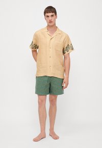 Camicia beige a maniche corte con bottoni, decorata con motivi ricamati verdi sulle maniche, abbinata a pantaloni corti a quadri verdi e blu.