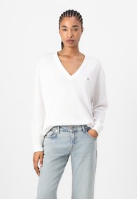 Pull à col en V blanc avec des manches longues, fabriqué en tissu doux. Porté avec un jean taille haute bleu clair, comportant un petit logo.
